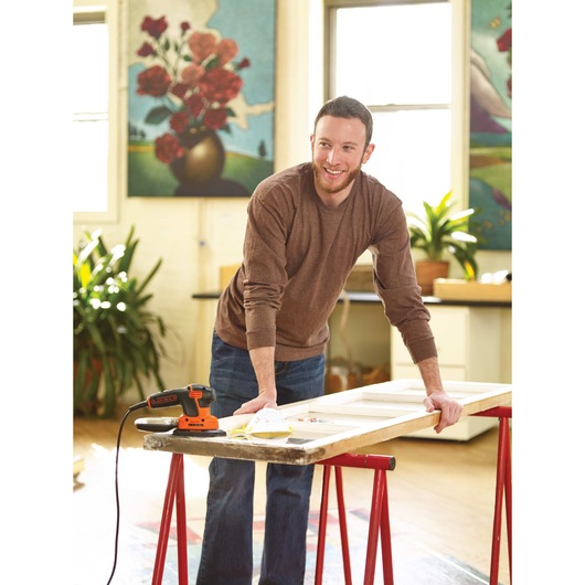 MOUSE Detail Sander at rest on wooden door placed on working stand besides a person in an indoor environment.