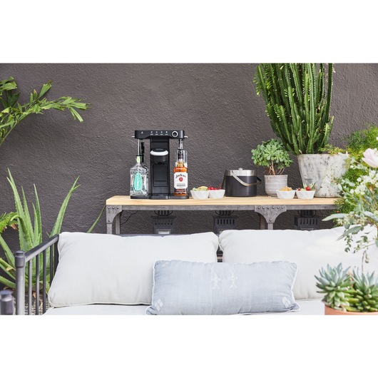 bev by BLACK+DECKER™ cocktail maker on an outdoor wooden serving table with an ice bucket and assorted garnishes